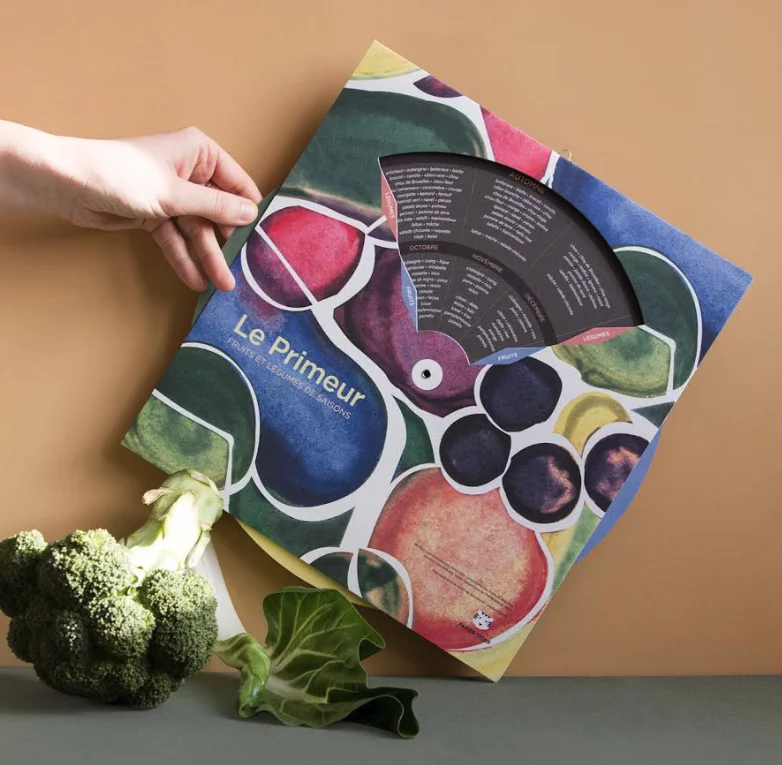 Calendrier perpétuel « Le Primeur » des fruits et légumes de saison, illustré d'aquarelles colorées et muni d'une roue tournante, tenu à la main devant un brocoli et une feuille verte.
